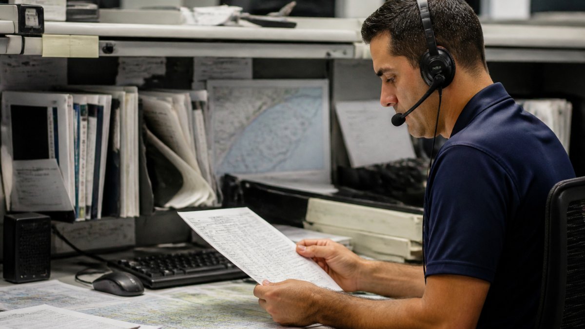 Aircraft dispatcher at a desk reviewing a printed flight plan with weather charts on a monitor, headset on, focused expression in a flight operations center