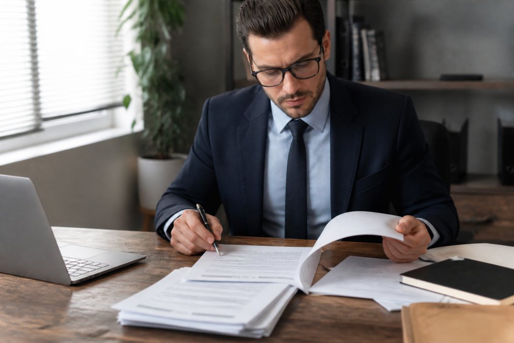 Business professional in formal attire reviewing detailed compensation contract documents and benefits packages at a wooden desk, with laptop and pen in hand, papers organized in front