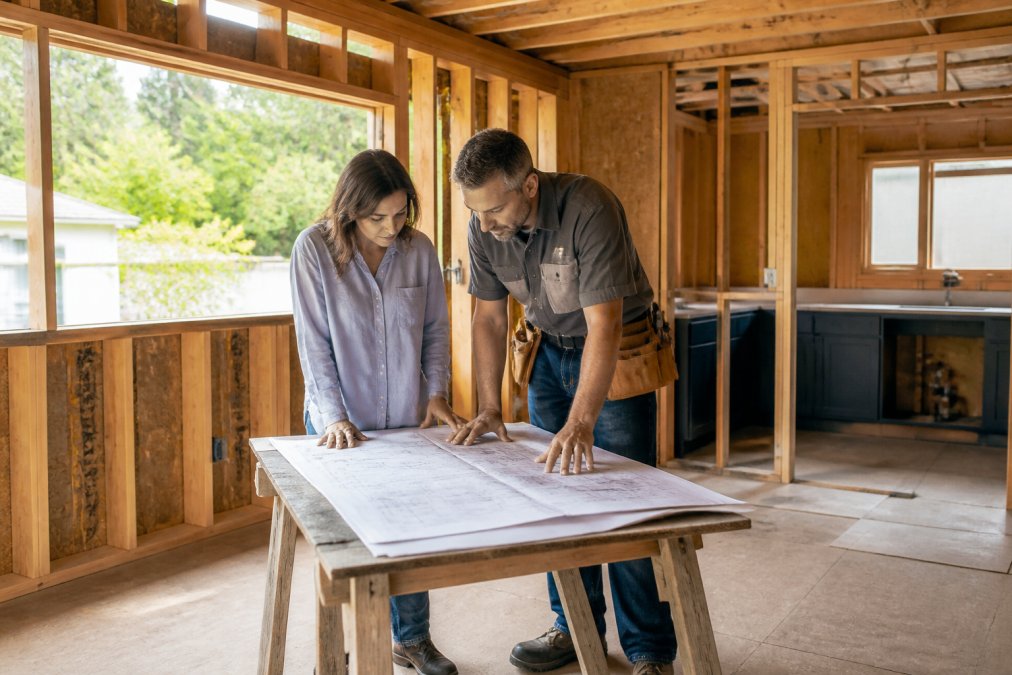 A homeowner and a contractor standing in an unfinished kitchen addition, blueprints spread across a sawhorse table, natural light through open framing
