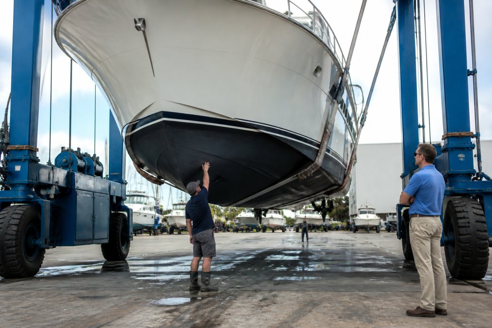 A yacht suspended in a travel lift at a boatyard, with a marine surveyor in work clothes inspecting the hull below the waterline while the owner watches from a safe distance