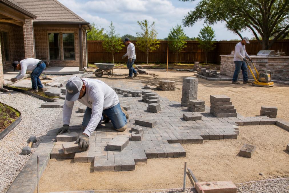 Professional hardscape crew operating equipment and installing pavers on a residential backyard hardscape project during daytime with materials and tools visible