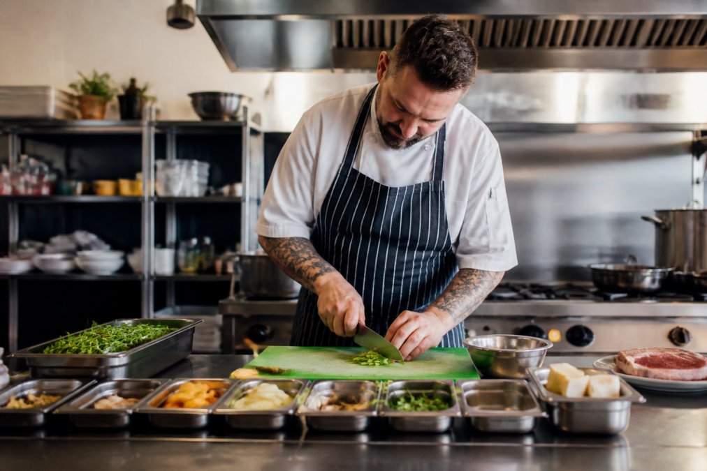 Professional head chef demonstrating precision knife technique while preparing mise en place on a stainless steel prep station in a bright, modern professional kitchen
