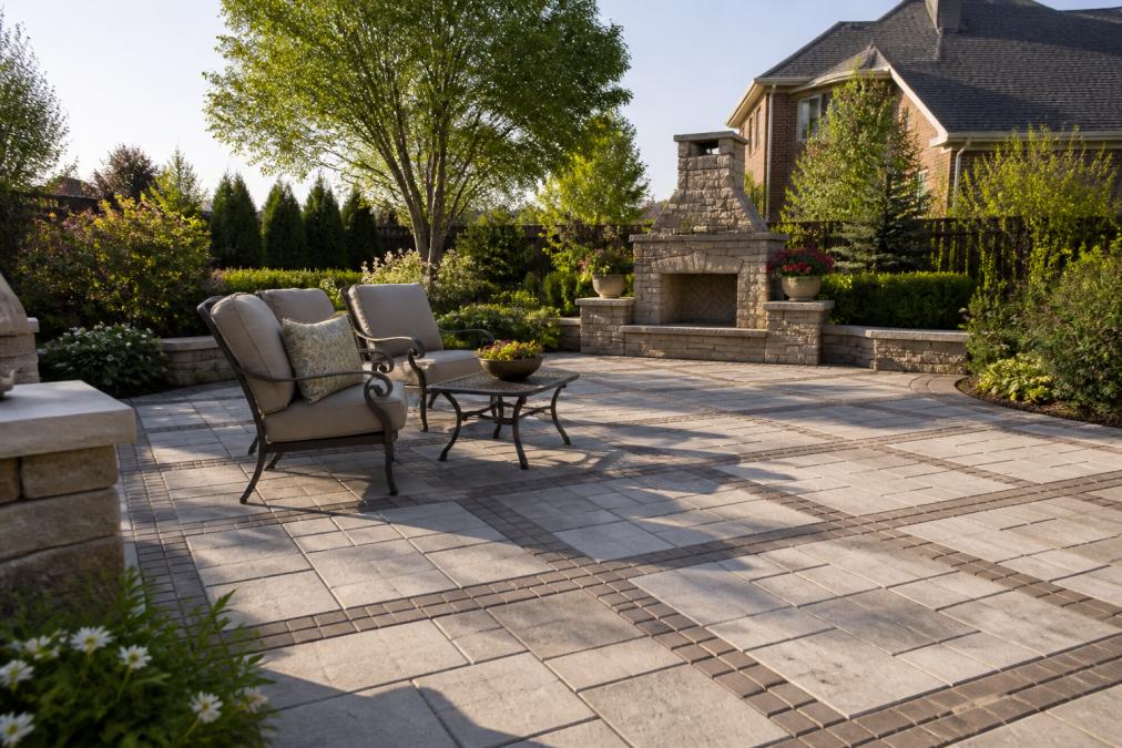 Finished stone patio with interlocking pavers creating geometric patterns, comfortable outdoor seating area with lounge chairs, mature landscaping and shrubs framing the space in a residential backyard during afternoon sunlight