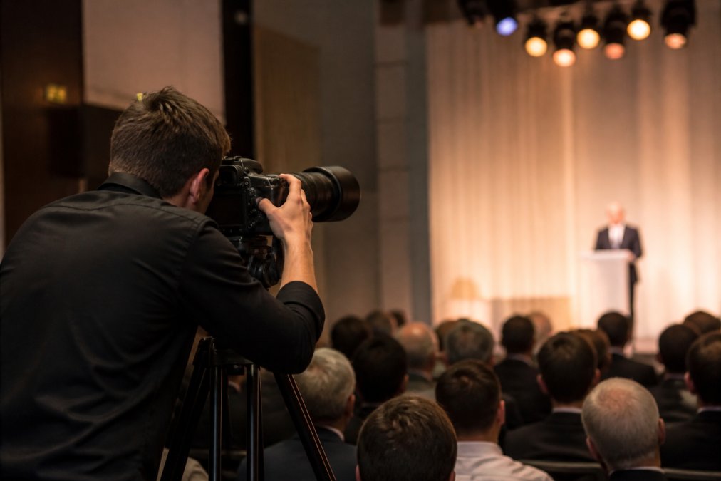 Professional photographer with a telephoto lens on a tripod photographing a corporate conference stage, audience heads in soft focus in the foreground, warm stage lighting overhead