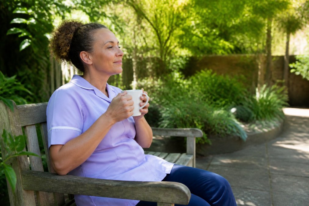 Compassionate carer taking a break in a peaceful garden, seated on a bench with natural daylight, appearing relaxed and supported with a cup of tea