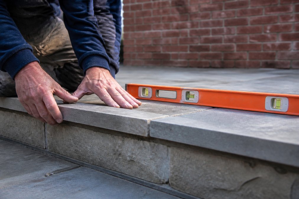 Close-up of a mason's hands running along a perfectly level bluestone patio edge, with a spirit level resting on the stone and a brick wall in soft focus behind
