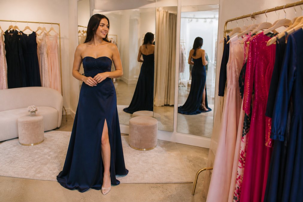 Woman in elegant formal dress trying on garments in a bright, welcoming boutique fitting room with soft lighting, full-length mirrors, and comfortable seating