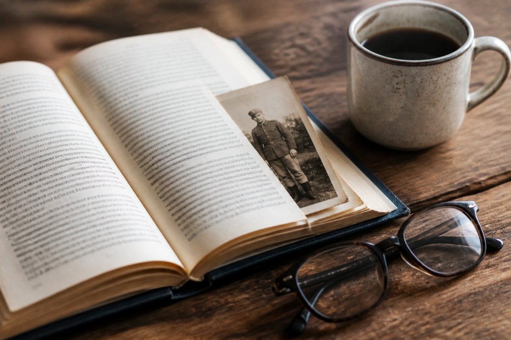 Open historical non fiction memoir with a vintage soldier photo, reading glasses, and coffee on a wooden desk