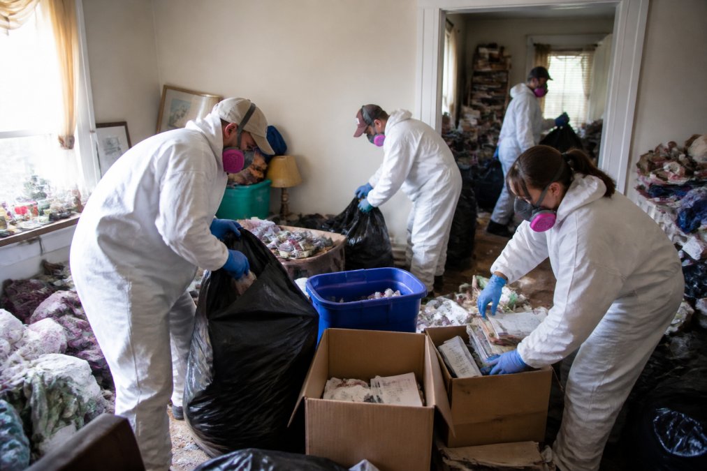 Professional team members in safety gear and protective equipment systematically sorting and removing items from a cluttered residential room during active cleanup, with boxes and bags organized by category