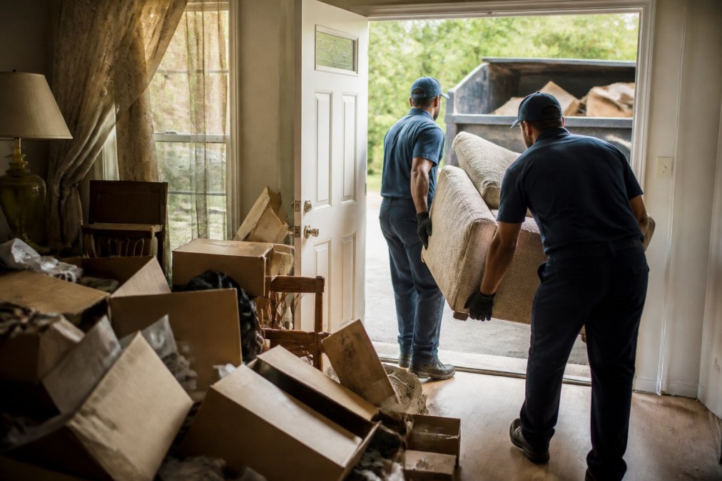 A team of two professional cleaners in uniform and gloves removing a large pile of old boxes and furniture from a residential living room into a large dumpster outside a bright, open front door