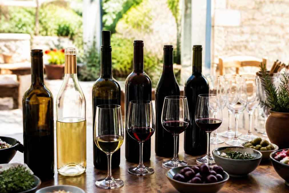 Eine Auswahl geöffneter Flaschen griechischen Weins mit unterschiedlichen Farben von Weiß- und Rotwein auf einem Holztisch, im Hintergrund Weingläser und mediterrane Zutaten wie Oliven und Kräuter