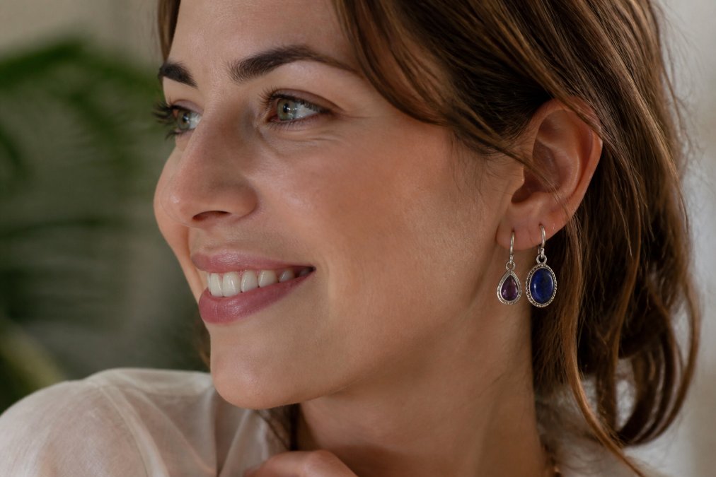 Nahaufnahme einer Frau mit strahlendem Lächeln, die verschiedene elegante 925er Silber-Ohrringe mit glänzenden Edelsteinen wie Amethyst und Lapislazuli trägt, natürliches Tageslicht, die Ohrringe funkeln subtil