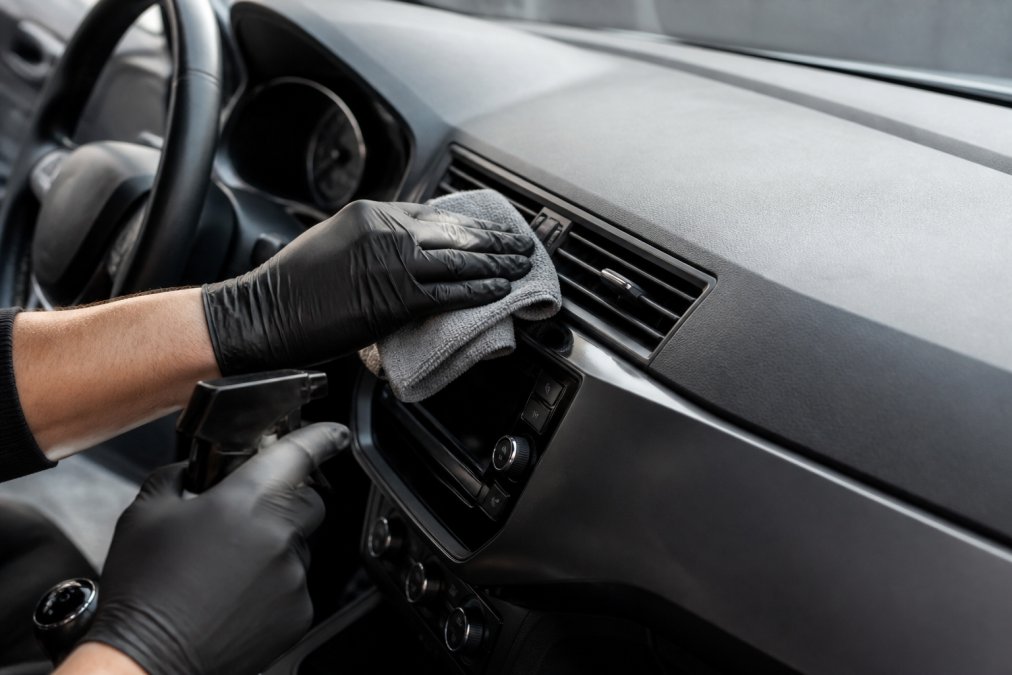Close-up van iemand die voorzichtig het dashboard reinigt met een microvezeldoek rond een touchscreen, met een fles interieurreiniger zichtbaar, focus op de voorzichtige beweging rond ventilatieopeningen
