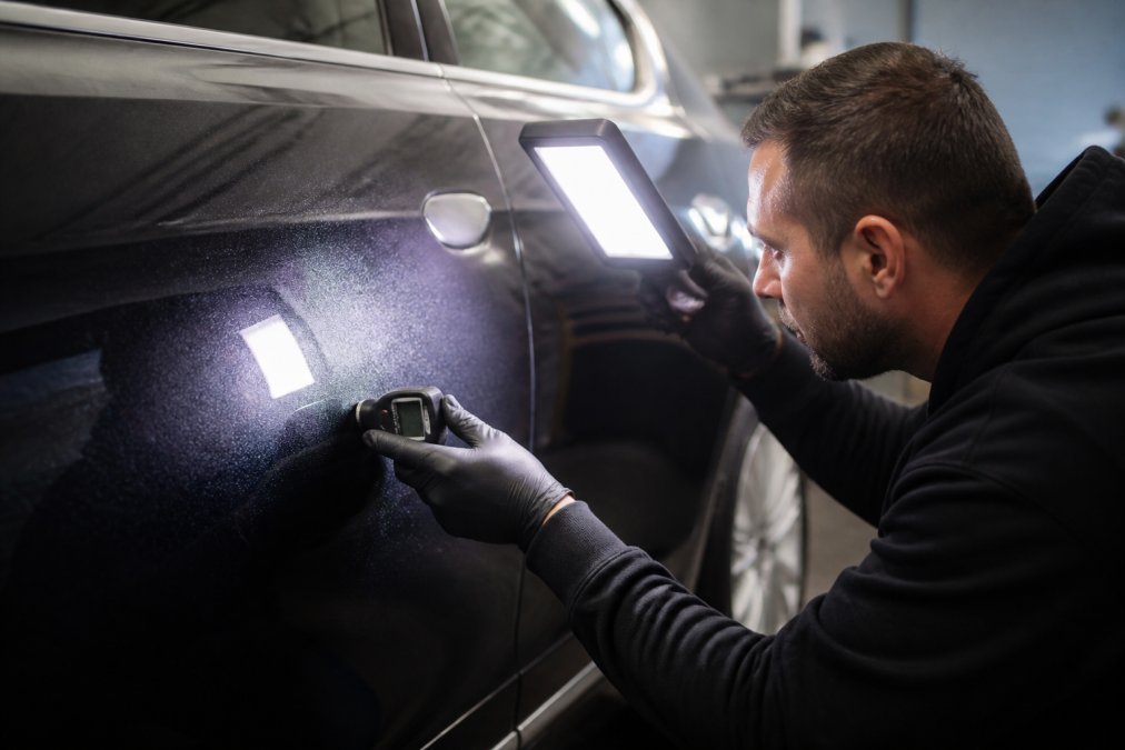 Close-up van iemand die met een inspectielamp de lak van een auto controleert in een garage, met een lakdiktemeter in de hand