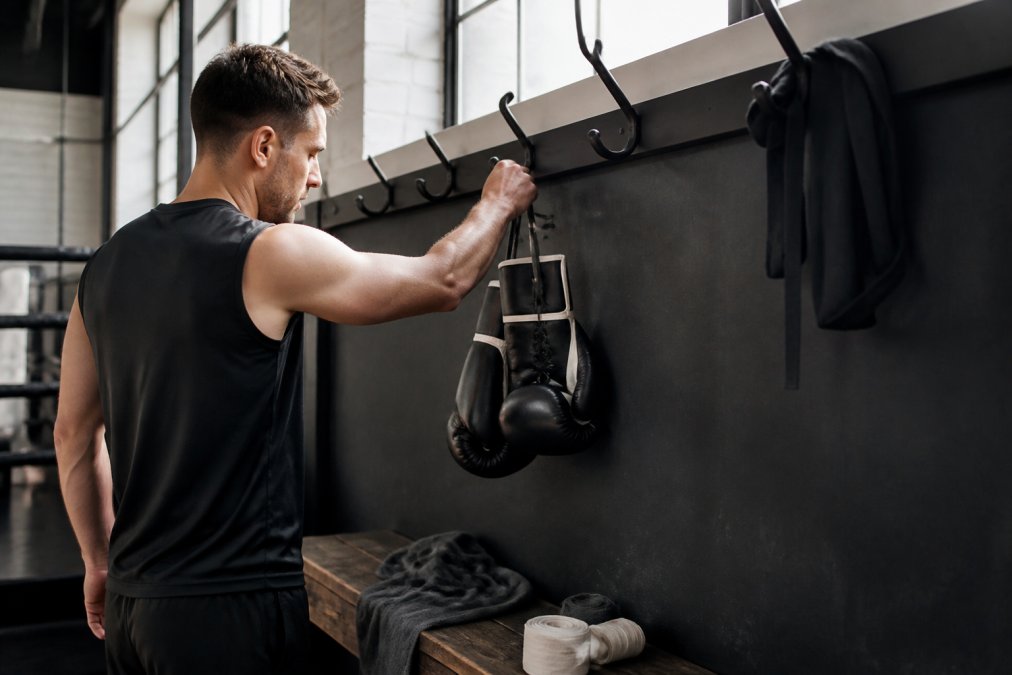 Een vechtsporter hangt zijn bokshandschoenen na een training aan een open haak in een goed geventileerde sportzaal, met zweetdoekjes en bandages op de achtergrond, natuurlijk licht door hoge ramen