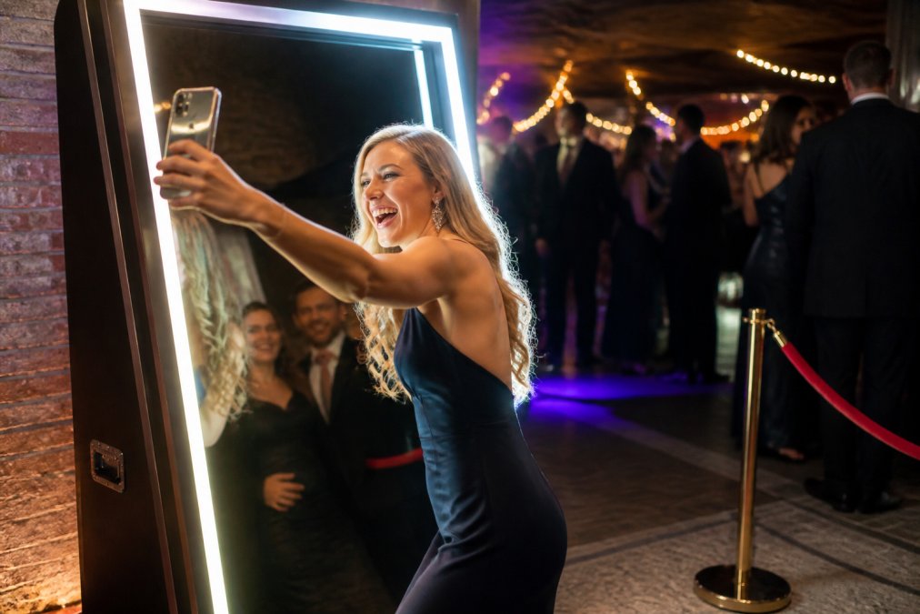 Een gast in avondkleding die lachend een selfie maakt bij een verlichte spiegelbooth op een feestlocatie, met op de achtergrond andere gasten en feestverlichting