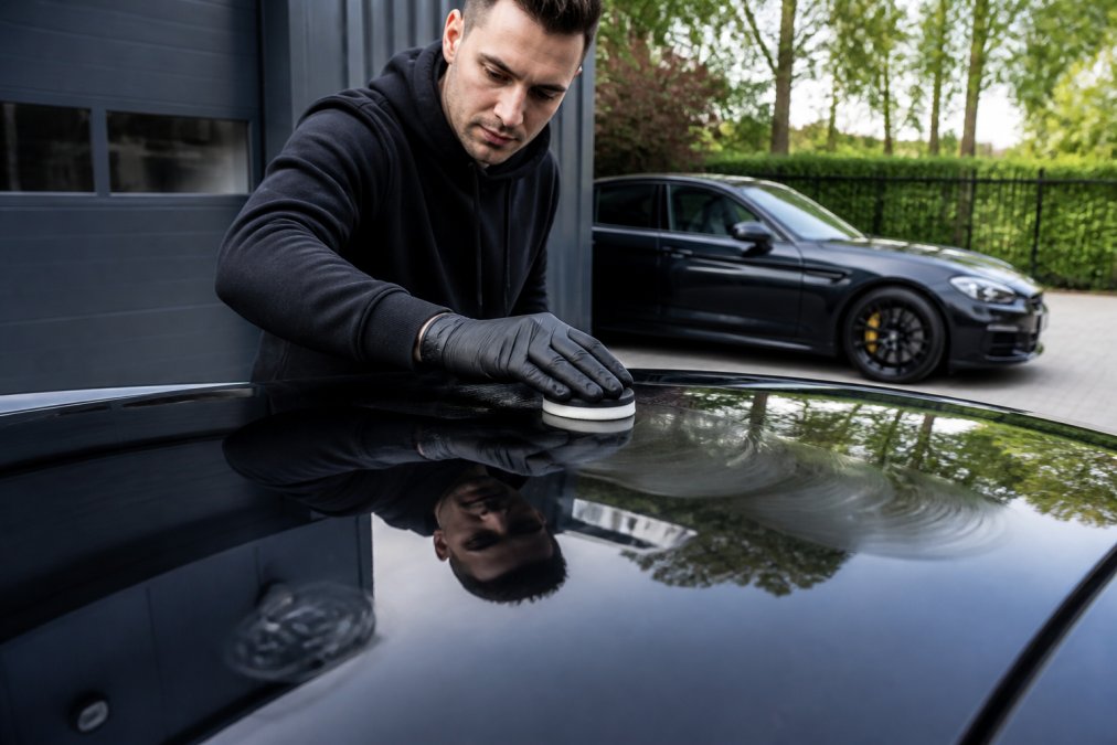 Professionele detailer brengt wax aan op een autodak met een schuimapplicatorpad, voorzichtige cirkelbewegingen, in helder daglicht met een schone auto op de achtergrond