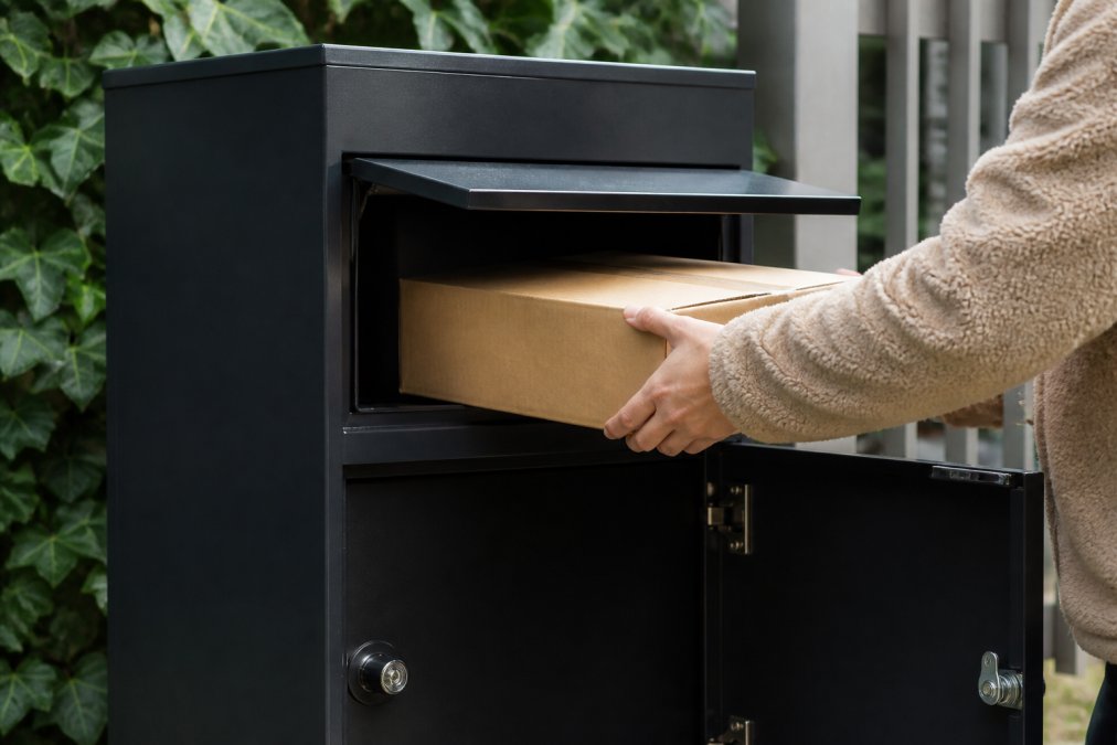 Close-up van een moderne pakketbrievenbus met zichtbare anti-hengelklep en versterkt slot, terwijl iemand voorzichtig een pakket in de veilige opening plaatst in natuurlijk daglicht