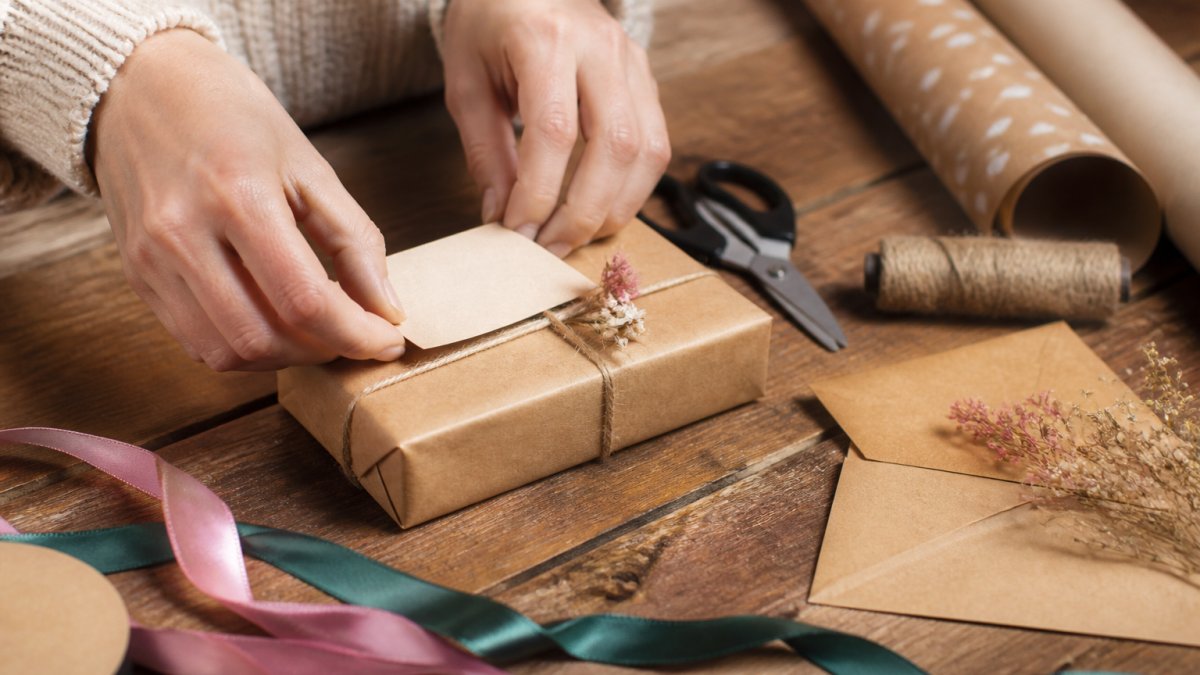 Handen die een klein cadeaudoosje inpakken met cadeaupapier en een kaartje, op een houten tafel met lint en schaar