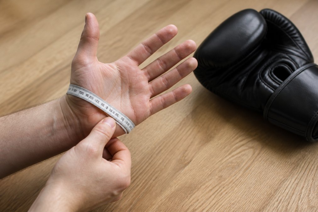 Close-up van iemand die een meetlint strak om de open handpalm wikkelt, gemeten vanaf de knokkels tot aan de pols, op een lichte houten tafel met een zwarte bokshandschoen ernaast