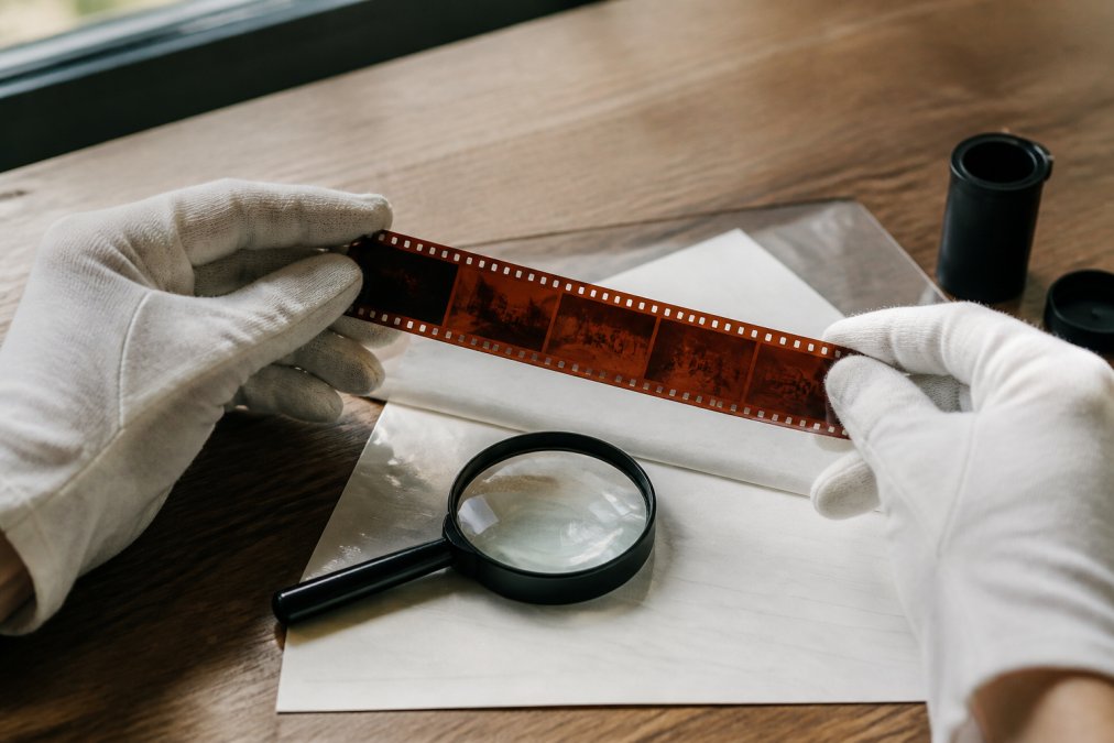 White-gloved hands inspect 35mm negatives against daylight, with a magnifying glass on the table.