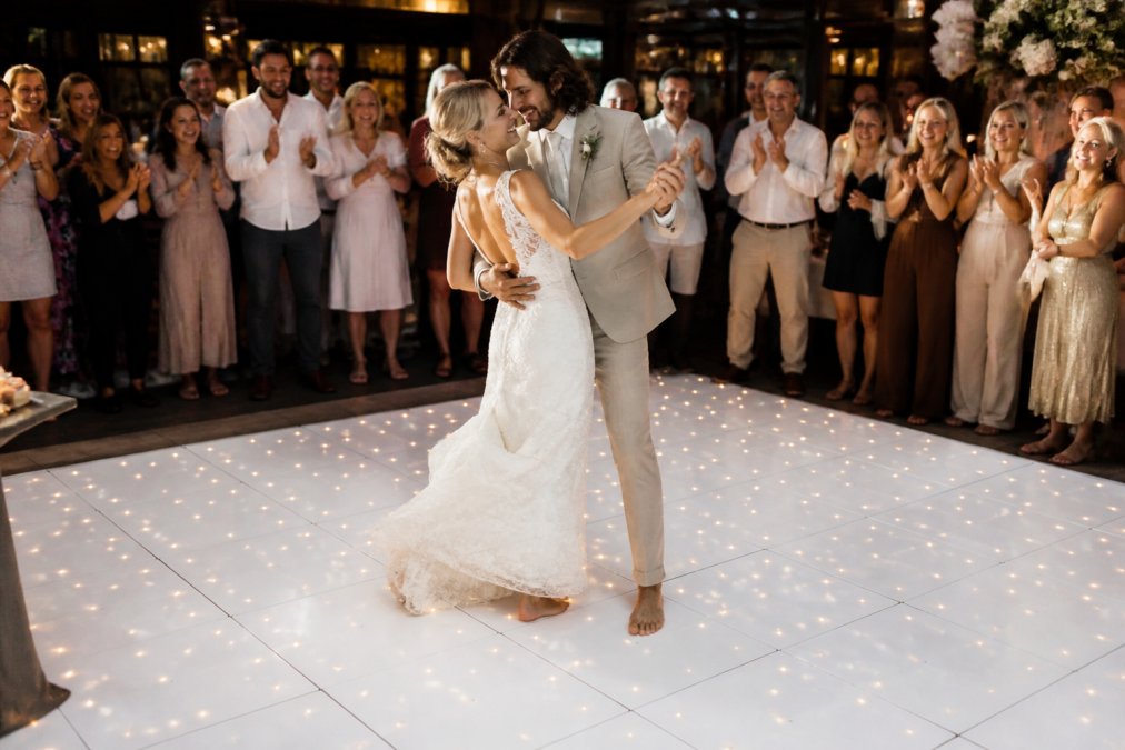 A couple in wedding attire dancing barefoot on a glowing LED dance floor while guests in evening wear clap and smile around the edges, warm ambient lighting