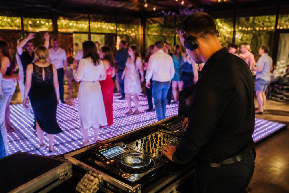 Professionele DJ achter een mengpaneel met koptelefoon, staande naast een verlichte dansvloer met dansende gasten op de achtergrond in een feestlocatie