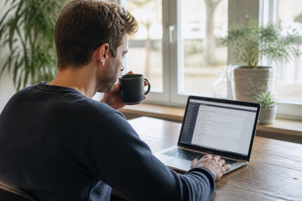 Iemand zittend aan een bureau met laptop en koffie, werkend aan website-onderhoud in een rustige thuisomgeving met natuurlijk licht