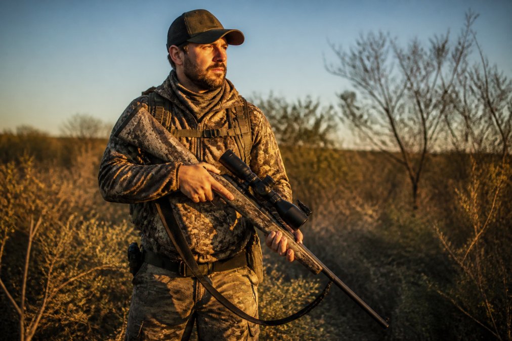 Hunter wearing layered camouflage apparel and holding rifle with thermal scope mounted, standing in brushy terrain at golden hour light