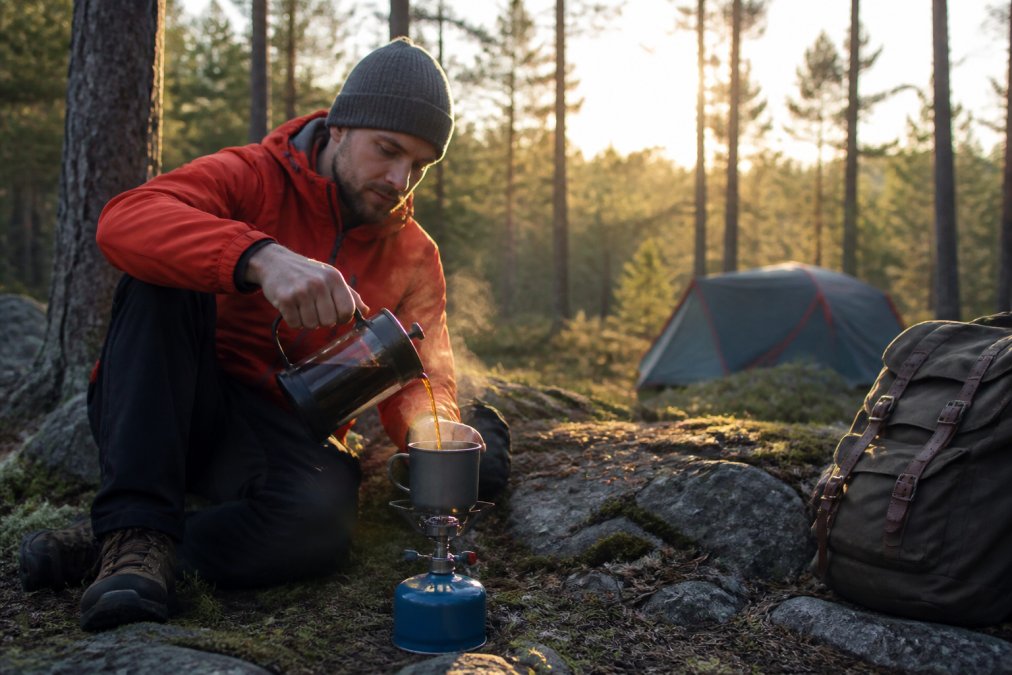 Hiker brewing coffee outdoors using manual coffee press with camping stove, steam rising from cup, forest camp setting with tent visible in background, morning light filtering through trees