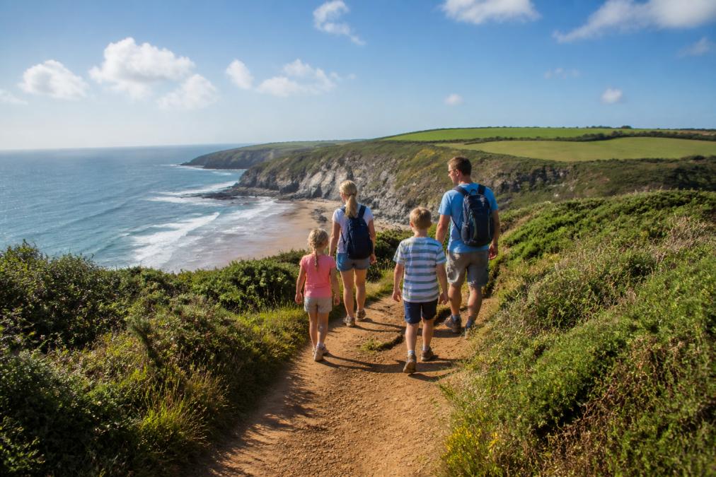 Children and adults walking together on a coastal path with dramatic cliffs and beach visible, clear sunny weather, family enjoying outdoor activity