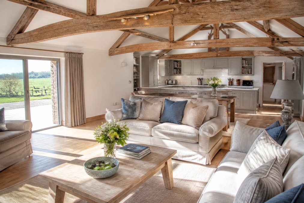 A bright, open-plan living area in a converted barn with comfortable sofas, wooden beams overhead, and sunlight streaming through patio doors onto a well-equipped kitchen beyond