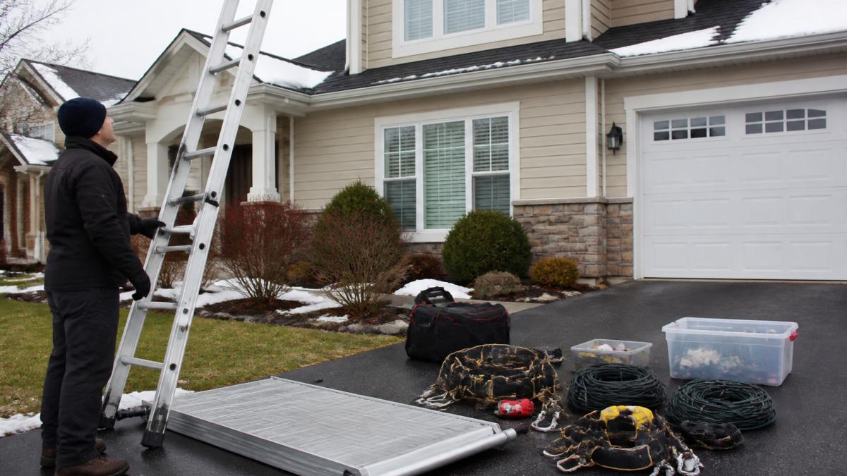 A homeowner in cold-weather gear standing at the base of a two-story house, checking a tall extension ladder and a roof platform laid out on the driveway before climbing