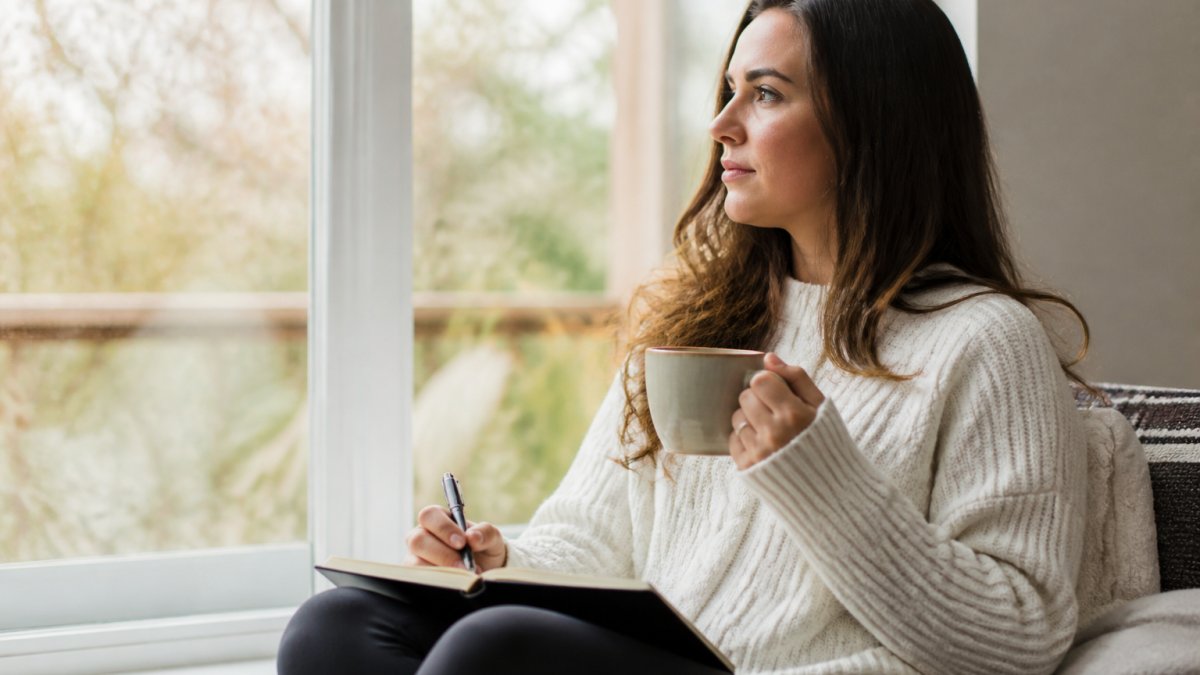 A woman sitting quietly by a window with a journal and a warm cup of tea, soft natural light, a calm and reflective mood suggesting a moment of emotional check-in