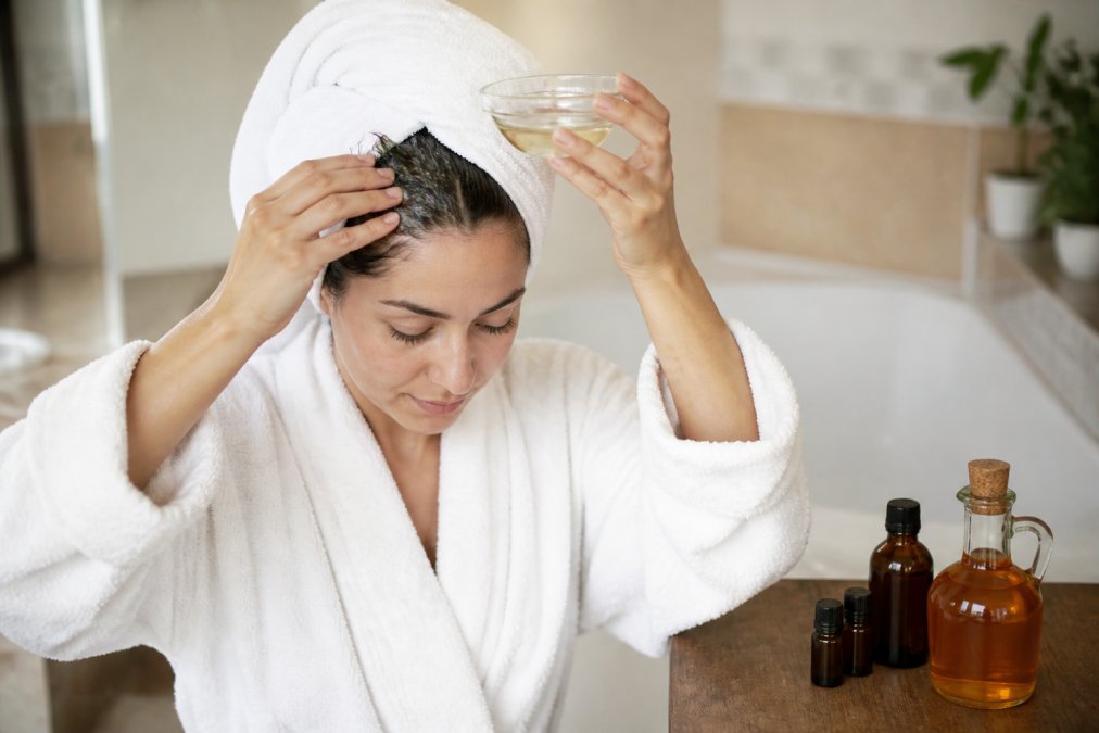 A woman with a towel-wrapped head gently massaging a small bowl of coconut oil into her scalp in a bright bathroom, with bottles of apple cider vinegar and essential oils on the counter