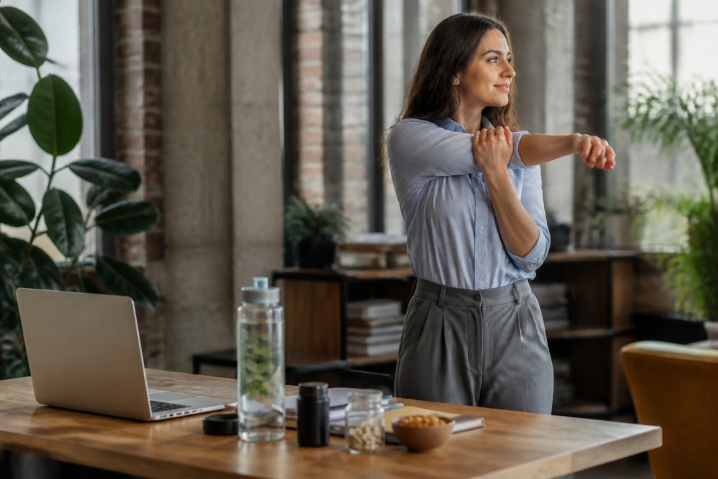 Professional at standing desk performing a quick shoulder stretch during work hours, with water bottle and wellness products on desk, natural office lighting