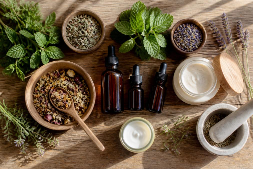 Overhead flat-lay of wellness products including glass amber bottles of herbal tinctures, loose leaf tea in wooden bowls, ceramic skincare jars, and fresh green herbs arranged on a natural wood surface with soft morning light