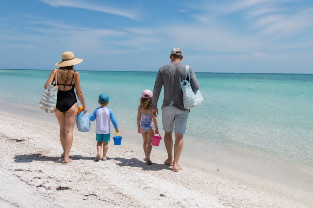 A family with young children walking along the sandy shoreline of Holmes Beach at mid-morning, carrying beach gear and looking out toward the calm, clear water of the Gulf of Mexico