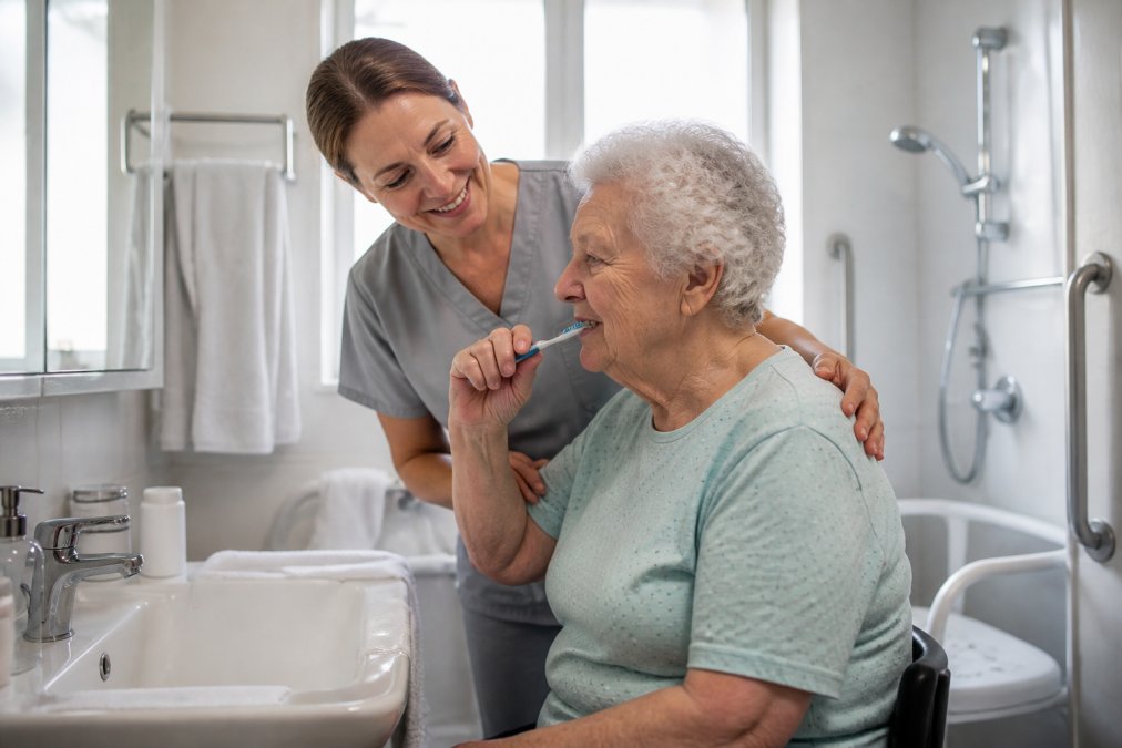 Professional caregiver assisting older adult with morning grooming routine in bright, accessible bathroom with grab bars and safety features visible
