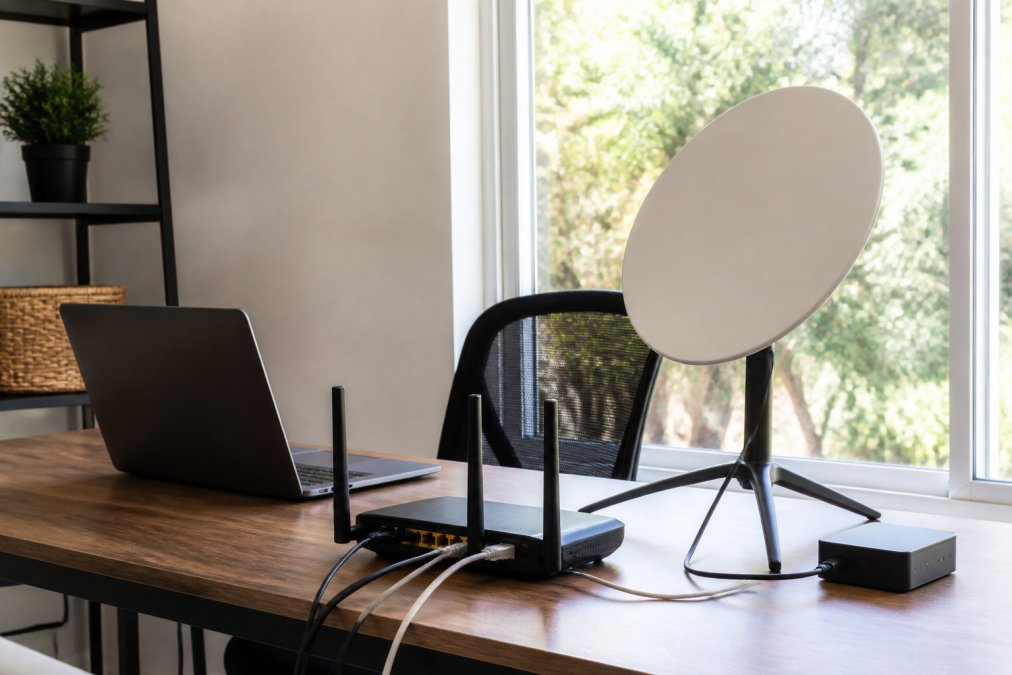 A clean home office desk setup showing a compact satellite dish on a stand near a window, with a dual-WAN router and a laptop on the desk, natural daylight streaming in