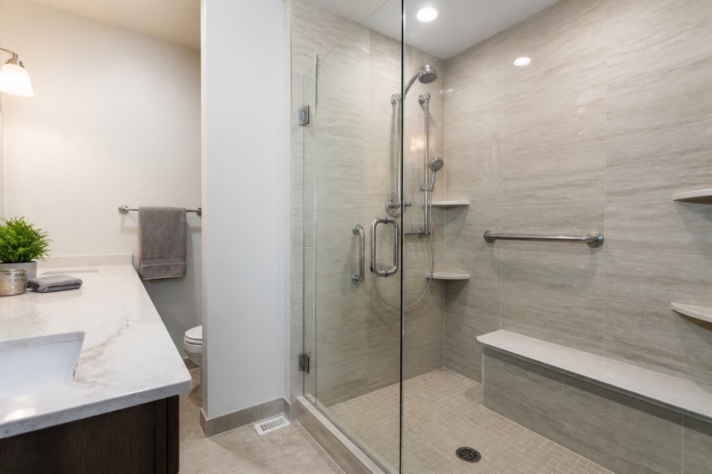 A clean, updated bathroom with a walk-in tile shower featuring chrome grab bars, a frameless glass door, and an accessible layout, photographed under bright overhead lighting