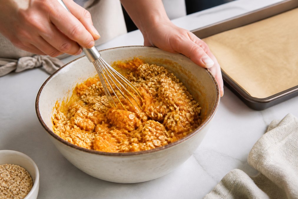 Close-up of hands whisking pumpkin puree and rolled oats together in a ceramic mixing bowl on a clean kitchen counter, with a baking sheet lined with parchment paper visible in the background