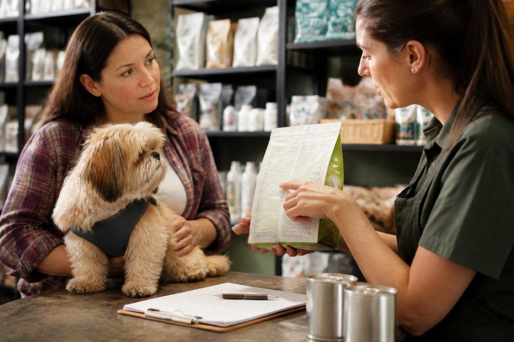 Pet owner discussing a dog's allergy symptoms with a knowledgeable boutique staff member, pointing to ingredient lists and discussing elimination diet options, with prescription documentation visible on counter