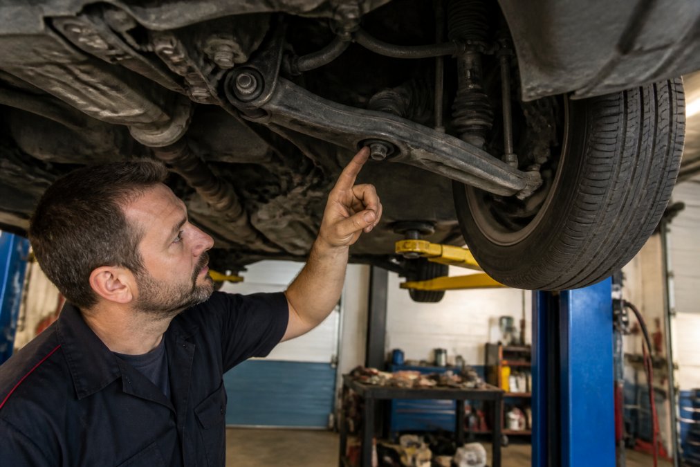 A mechanic in a clean independent repair shop pointing at a worn lower control arm bushing on a Honda Civic lifted on a two-post lift, with the undercarriage clearly visible