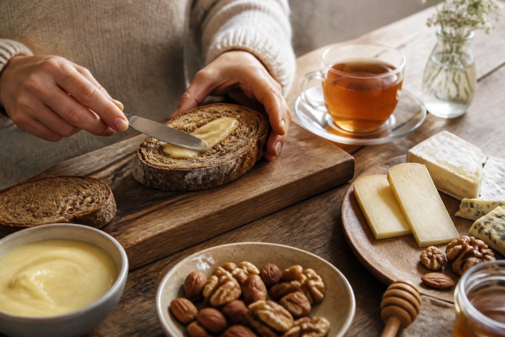 Hände, die cremigen Honig auf frischem dunklem Brot streichen, daneben Käsestücke, geröstete Nüsse und ein Glas Tee auf rustikalem Holzuntergrund mit natürlichem Licht