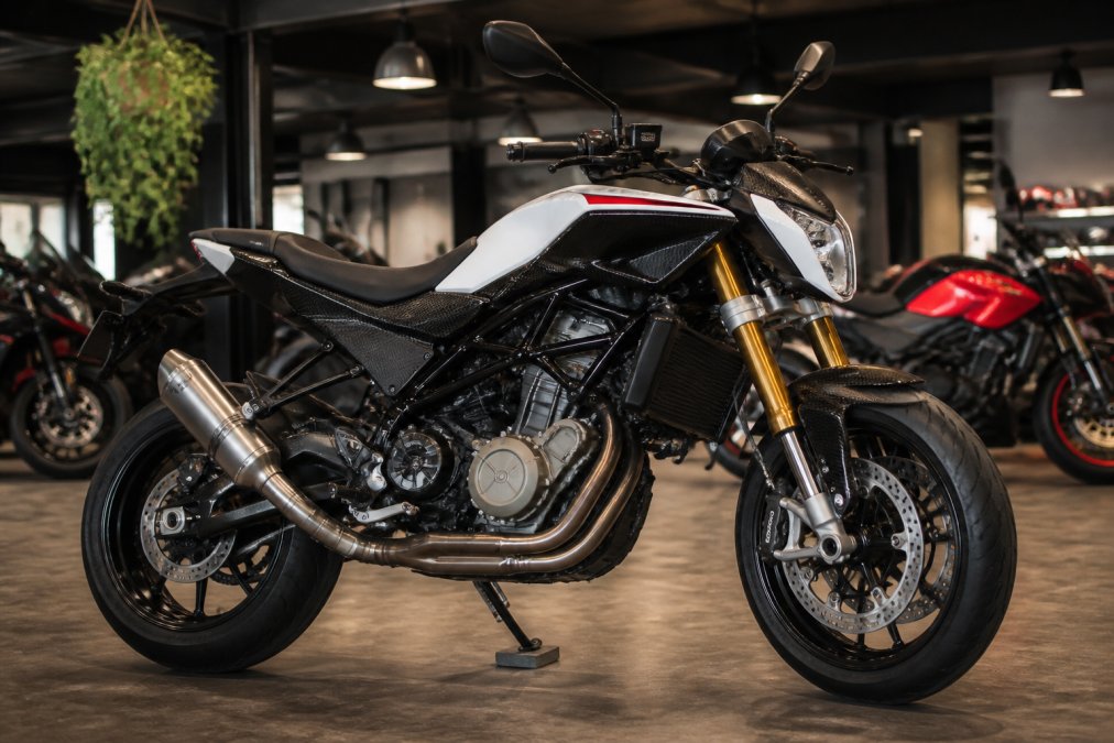 Husqvarna Nuda 900R in showroom-omgeving, detail van carbon kappenset, Öhlins-vering en titanium uitlaat zichtbaar, professionele werkplaatsverlichting