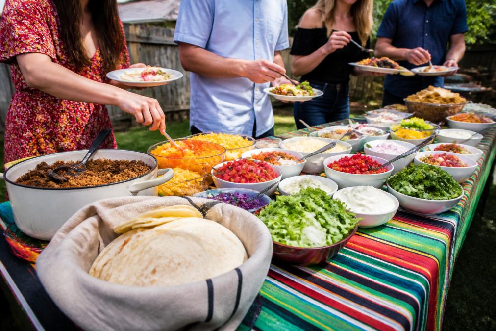Guests building plates at a casual outdoor budget taco bar with colorful toppings and warm tortillas.