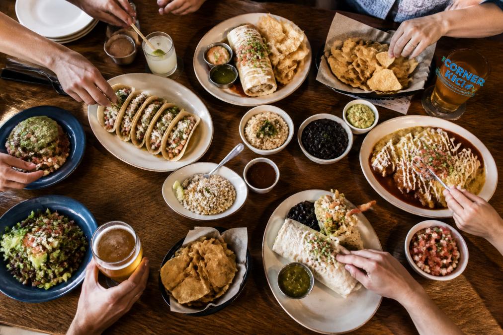 Overhead view of family-style dishes spread across a wooden table, scratch-made tacos, signature burritos, and shared plates of fresh ingredients, with multiple hands reaching across to serve themselves, warm ambient lighting
