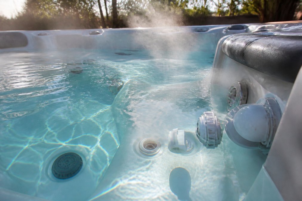 Close-up of pristine hot tub water with visible steam rising, showing the injector system and check valve components, crystal clarity revealing the tub floor with natural sunlight reflecting off the surface