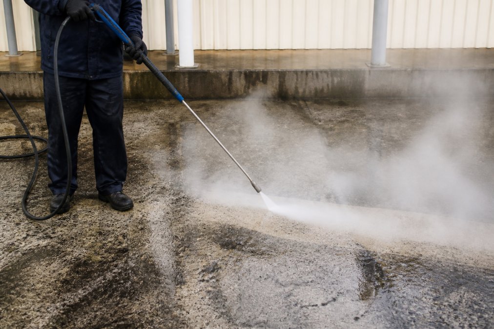 Professional demonstrating hot water pressure washer spray technique on concrete driveway with visible oil stains, showing steam and cleaning action in progress
